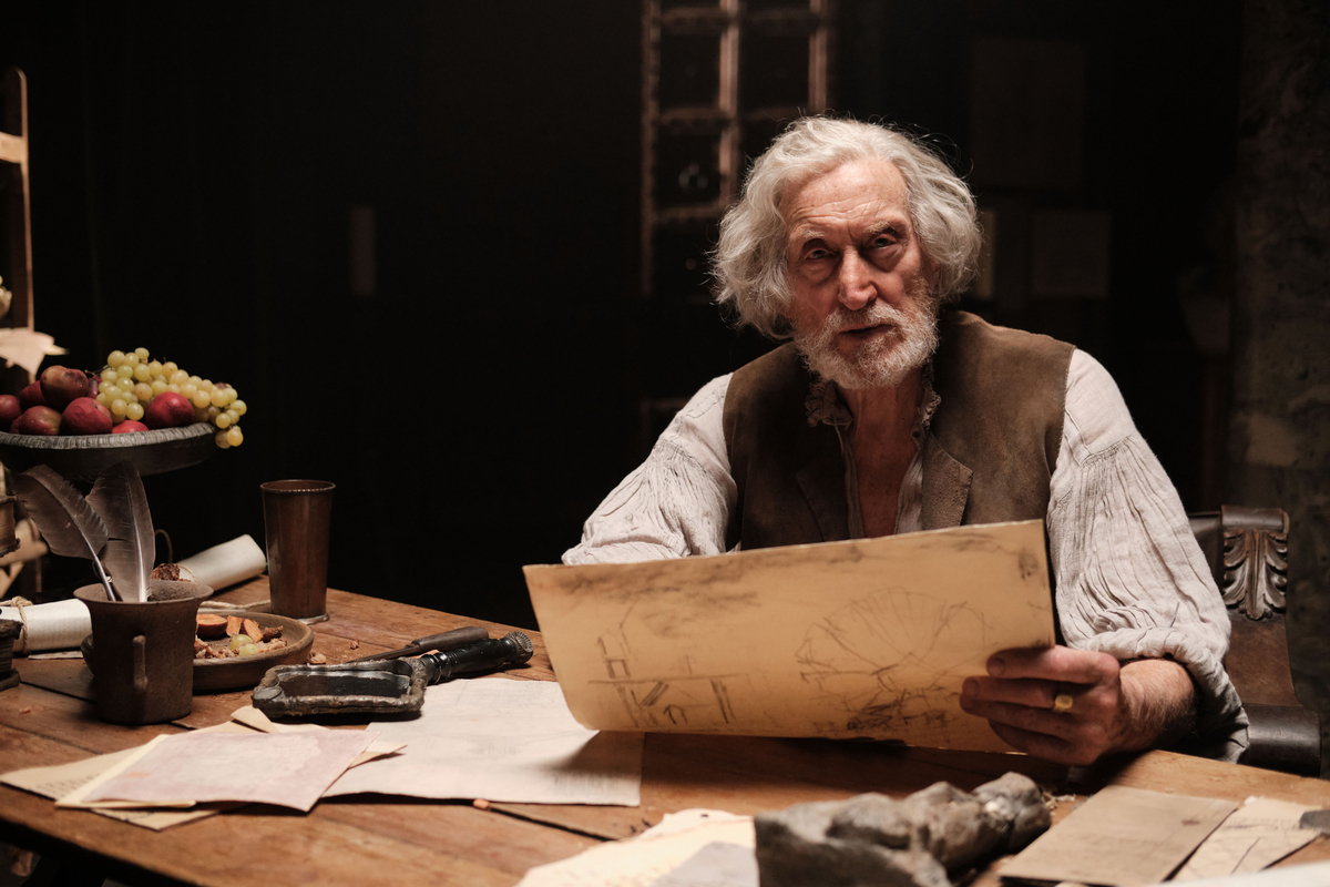 Charles Dance as older Michelangelo, he has shoulder length white hair, a beard and is in his late 70s. He sits at a wooden table with plans around him on orange coloured paper, to his left is a fruit bowl with apples and grapes.