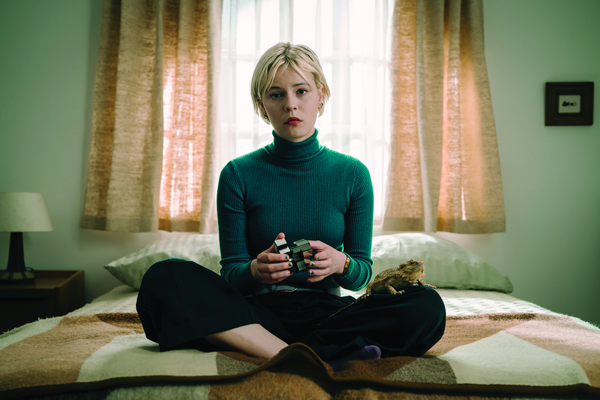 A white woman in her 20s wearing a green polo neck top sits on a bed playing with a Rubik's cube