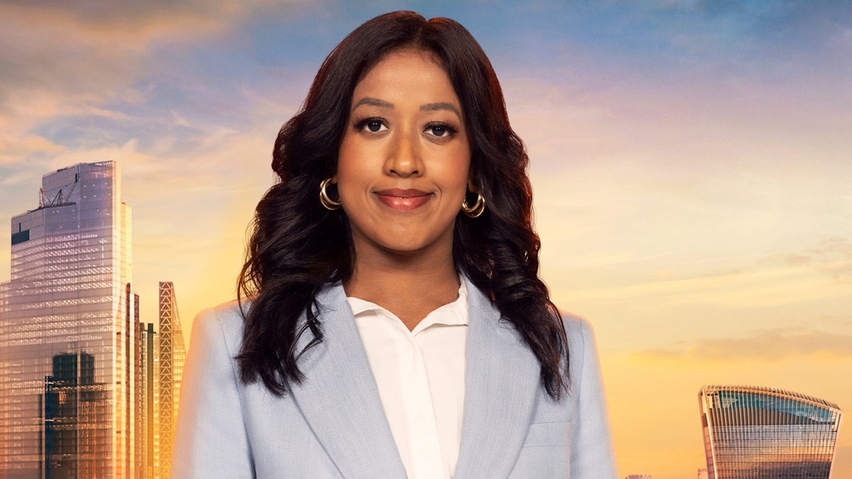 Anisa Khan looks into the camera in front of the London skyline, wearing a grey-blue blazer and white shirt