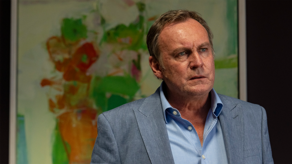 Philip Glenister, a white man in his sixties, stands indoors in a suit, with out-of-focus blocks of green, red and orange (possibly a map) on a wall behind him