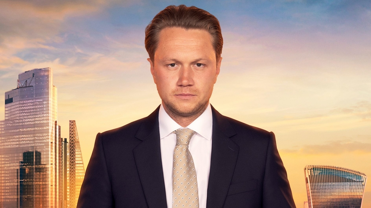 Dean Franklin looks into the camera in front of the London skyline, wearing a black blazer, white shirt and light brown tie with white and black diamonds