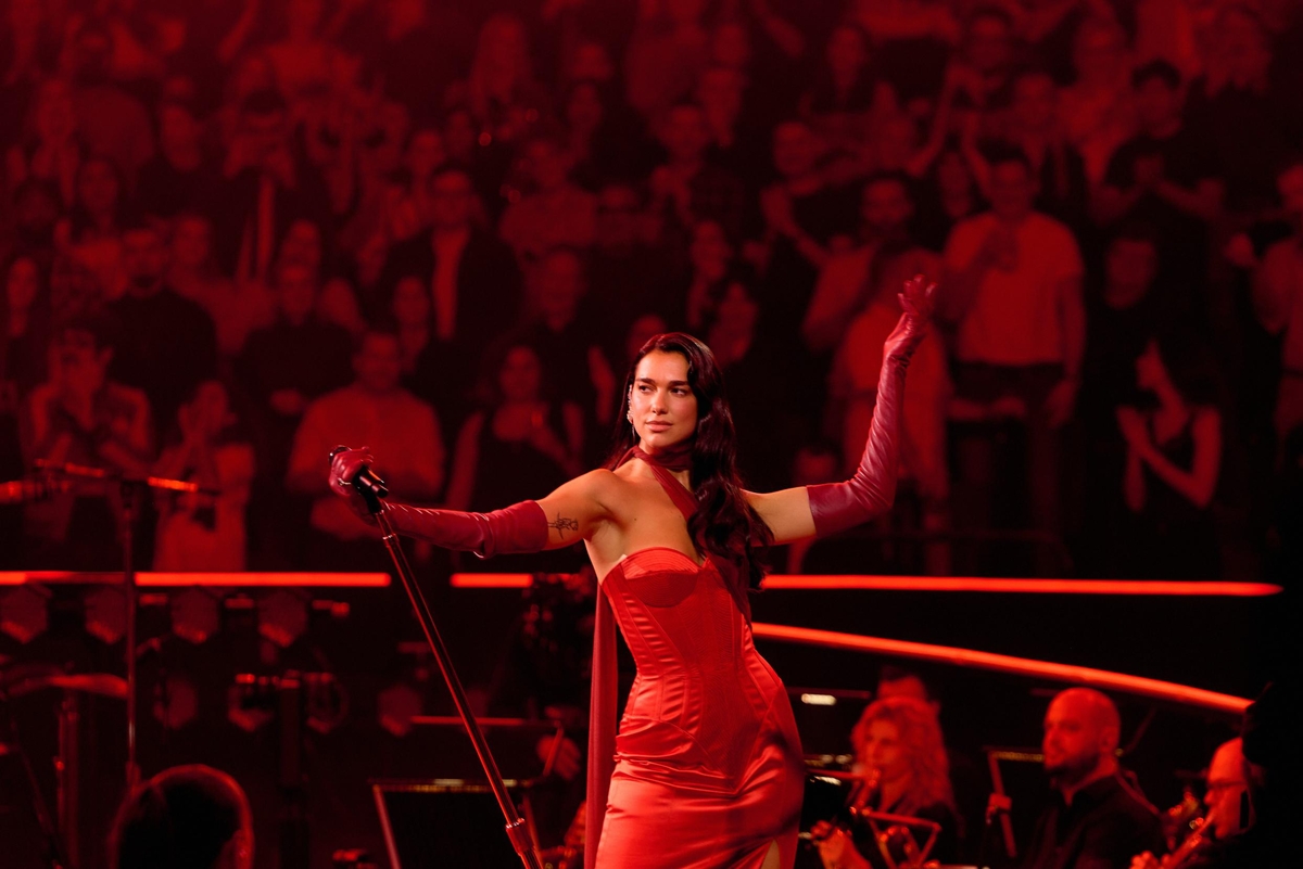 Dua Lipa, a woman in her late 20s, stands on a stage wearing a red corset dress, long red gloves, and holds a microphone away from her face. The audience is also bathed in a red light, and behind the stage there are a few members of the orchestra.