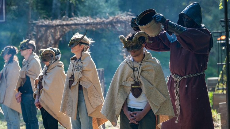 Five players stand in a line with capes and horned masks on, while the player furthest to the right has a bucket of black liquid poured over his head from behind by a big masked man