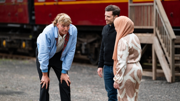 Two white men and a woman wearing a headscarf stand by a train looking regretful