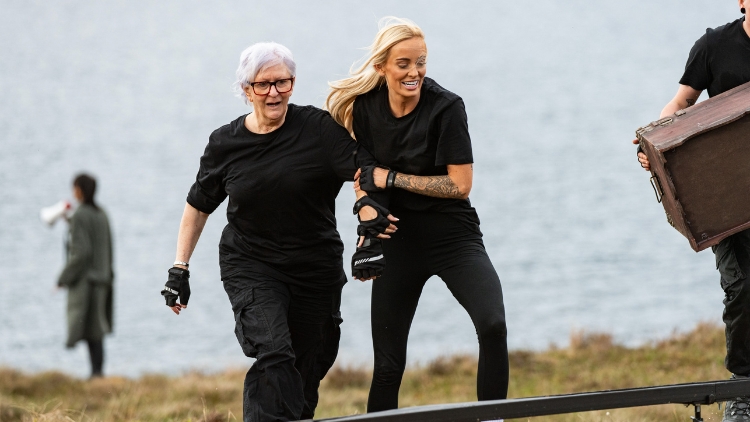 A white woman wearing all black helps an older white woman also wearing all black up a hill while smiling