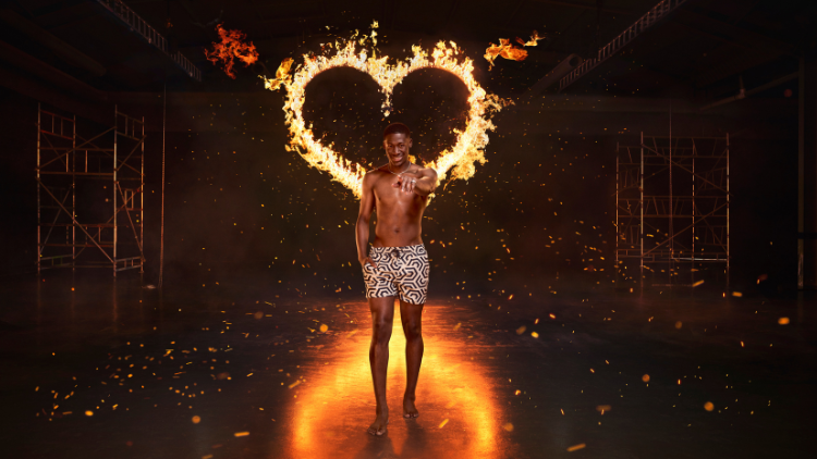 Ayo Odukoya stands in front of a heart that has been set on fire, against a black backdrop