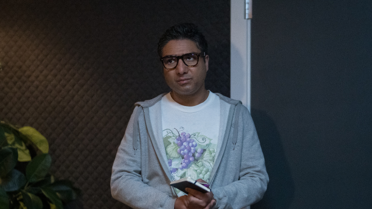 Nick Mohammed stands indoors, wearing large-framed glasses, looking annoyed