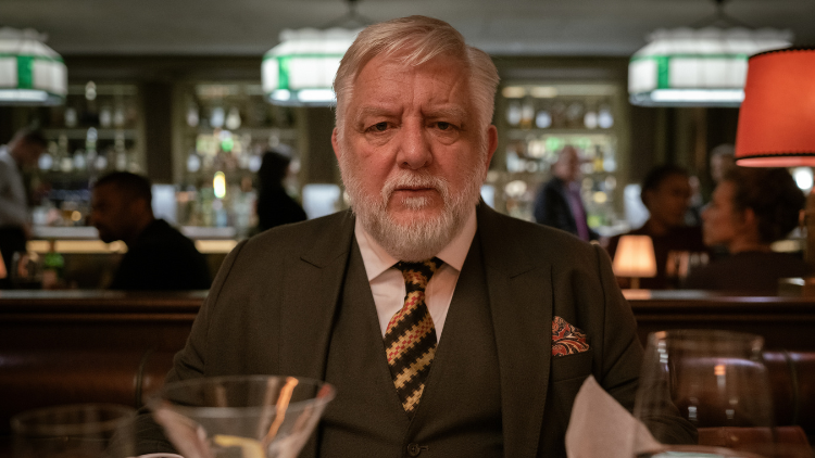 Simon Russell Beale sits in a restaurant, wearing a suit and looking serious