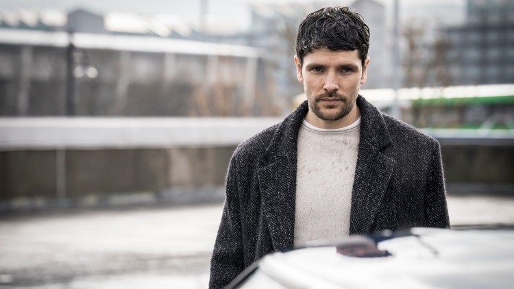 Colin Morgan stands by a car, head tilted down slightly, looking serious
