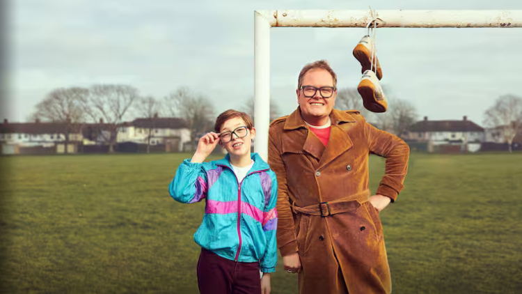 Alan Carr and Oliver Savell stand leaning on a football goalpost, a pair of shoes tied together and hanging from the crossbar