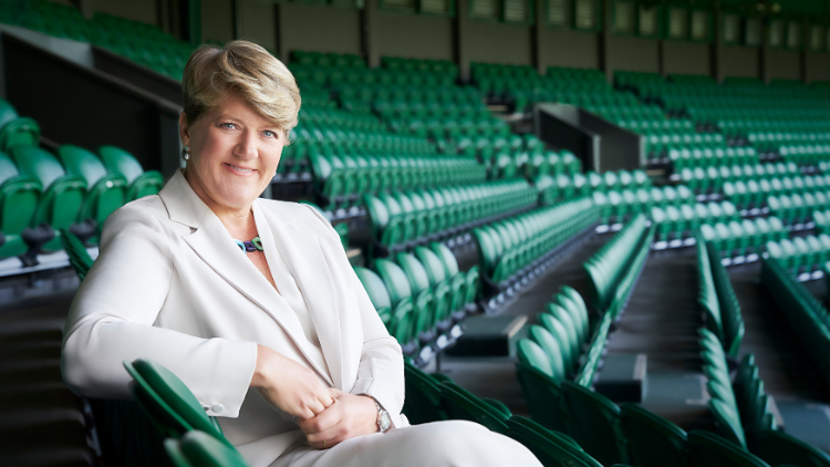 Clare Balding sits in an otherwise empty stadium at Wimbledon, looking into the camera and smiling 