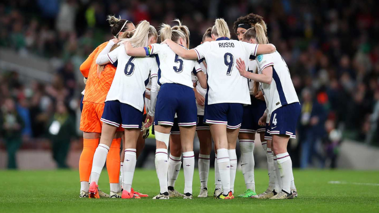 The England women's squad stand in a huddle on a pitch