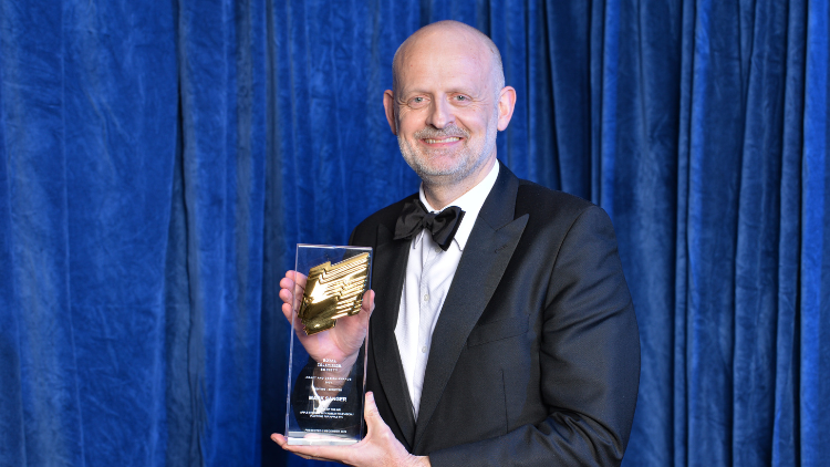 Mark Sanger, a white man in his fifties, smiles and holds an RTS award up to the camera in front of a blue backdrop, smiling in black tie