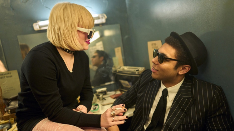 Eve Austin, a white woman in her twenties, and Levi Brown, a Black man in his twenties, smile at each other while speaking, both in sunglasses. Levi wears a hat and pinstripe suit and Eve a black choker and top