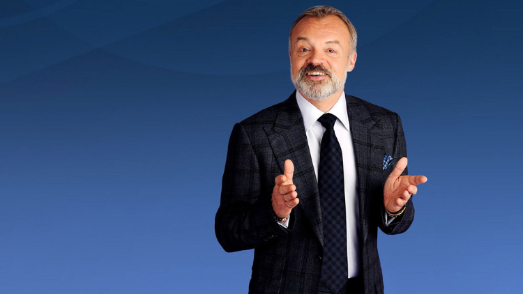 Graham Norton stands in front of a dark blue backdrop, gesturing with his hands and smiling, as if mid-conversation