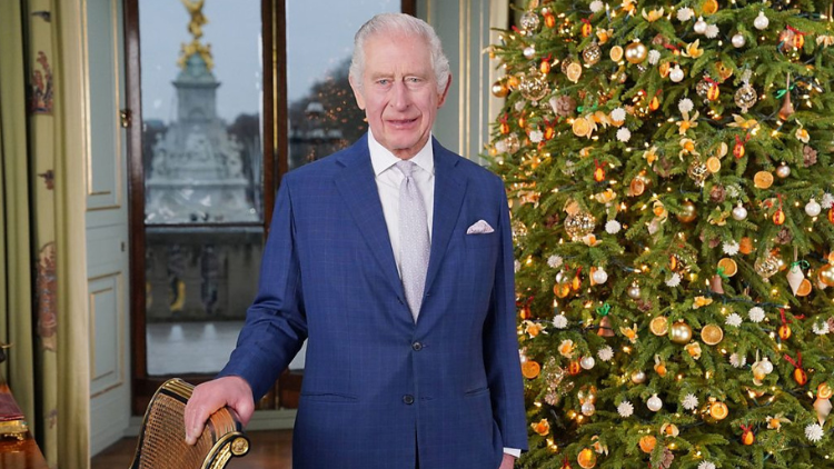 King Charles stands indoors, looking into the camera, one hand on the back of a chair next to him and a Christmas tree behind him