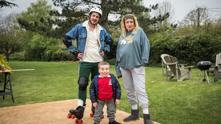 Dustin Demri Burns, Lenny Rush and Daisy May Cooper stand in a large outdoor garden, Burns wearing roller skates, a crash helmet, elbow pads, knee pads and wrist pads