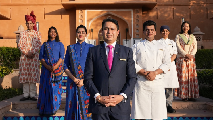 Seven people in formalwear, including two in chef whites, stand in front of a hotel in India