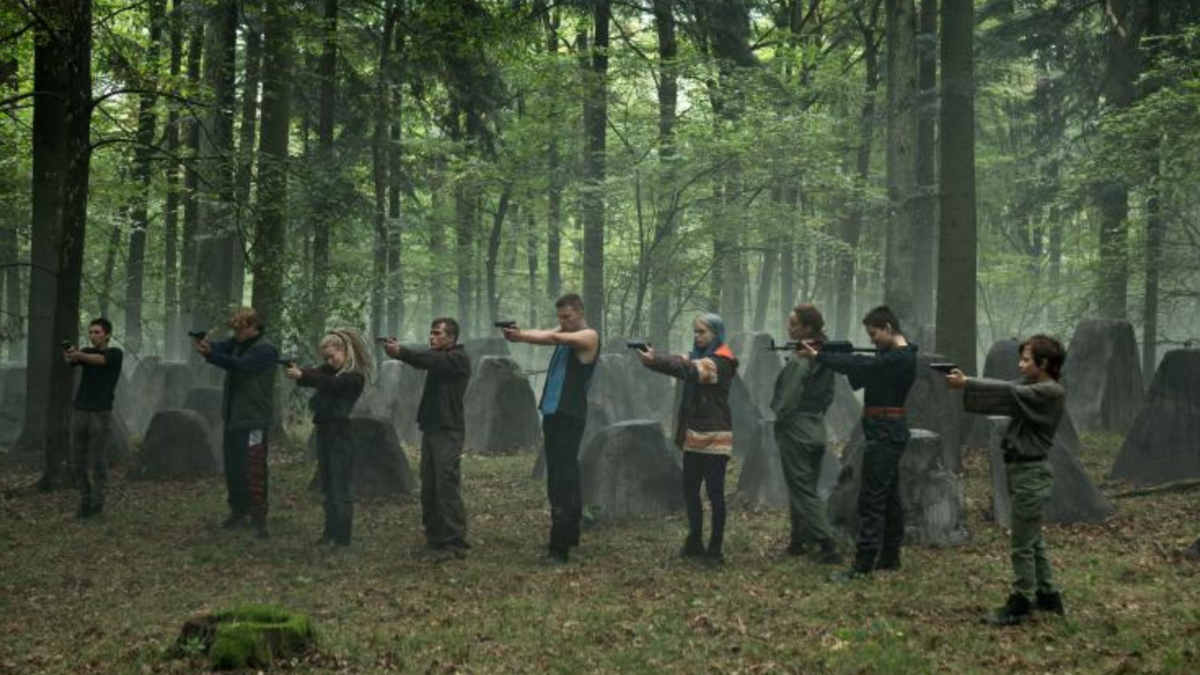 A group of nine people stand in a forest, aiming guns at the same target, which is off-screen