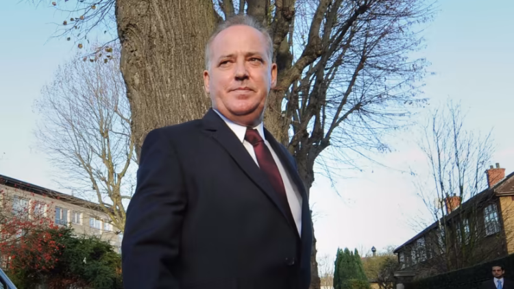 Michael Barrymore stands outside in a suit, in front of a tree and homes 