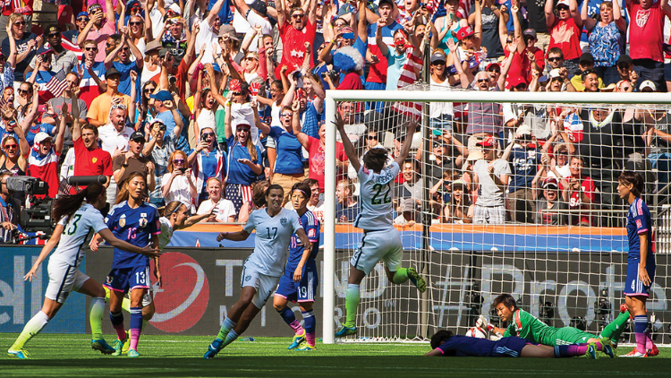 In a football stadium, American women's players cheer by the Japanese goalpost, with their opponents looking on, desolate. The players are in front of cheering America fans