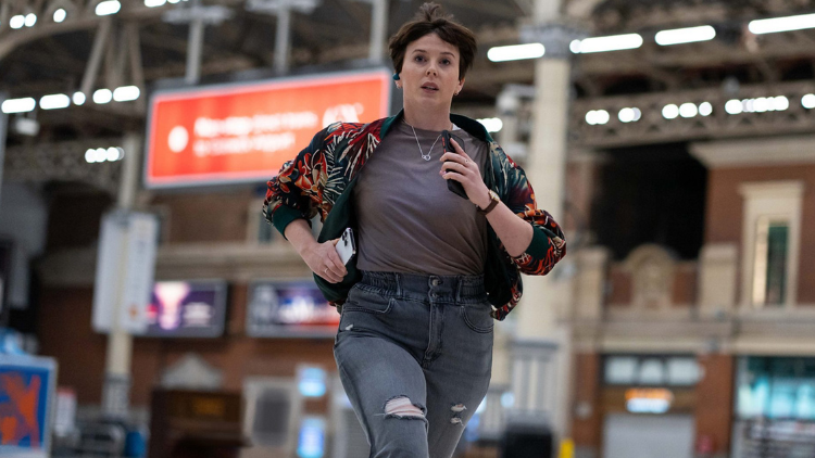 Alexandra Roach as Abby Aysgarth runs in a train station, holding two phones with what appears to be a small blue Bluetooth device in her ear, looking stressed