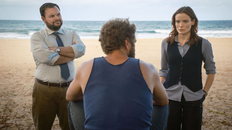 Lloyd Griffith and Anna Sampson stand in character on a beach, with the sea behind them. They are stood in front of a man who has his back to the camera and wears a navy tank top with curly hair. Griffith is in his early 40s and has dark hair and a beard. He wears a stripey shirt and tie, and khaki green trousers. Sampson wears a black waist coat and blue shirt and has curled brown hair. Both have a light skin tone. 