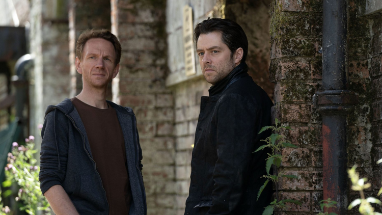 John and Michael Rebus stand by a house, looking into the middle distance, unsmiling