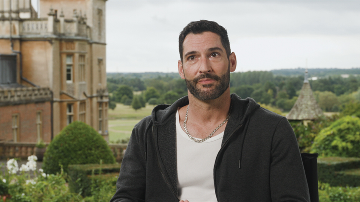 Tom Ellis sits in front of a pastoral landscape of hills and a church