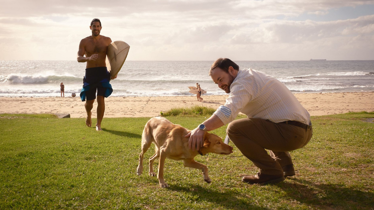 Lloyd Griffith leans down to stroke a golden lab on the beach, in character. He is in his early 40s and has dark hair and a beard. He wears a stripey shirt and tie, and khaki green trousers. A surfer runs towards him holding his board. He is smiling and wears long blue swim shorts. He has a medium skin tone and is in his mid 30s. This is Tai Hara. 