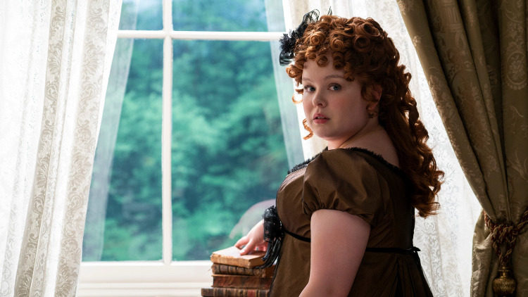 Nicola Coughlan as Penelope Featherington with her hand on a pile of books, looking over her shoulder at the camera 