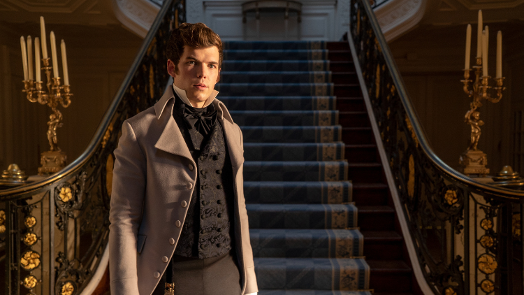 Luke Newton as Colin Bridgerton stands in front of a grand staircase