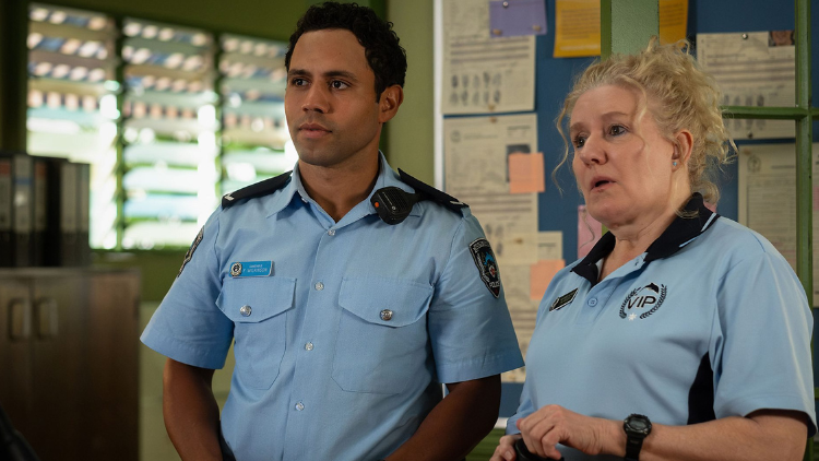 Aaron McGrath, and Celia Ireland are in character, wearing police uniform. She has blonde curled hair tied up in a bun, is in her late 50s, and has a light skin tone. He is in his late 20s, has a medium skin tone and black hair. They look at someone unseen off camera. 