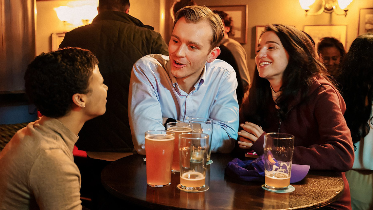 Myha’la Herrold, Harry Lawtey and Marisa Abela sit around a table in a pub, talking animatedly while smiling