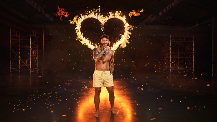 Sam Taylor stands in front of a heart that has been set on fire, against a black backdrop