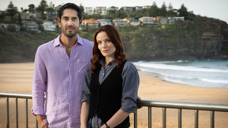 Tai Hara stands with Anna Samson in front of a beach. She is wearing a black waistcoat over a purple striped shirt. Her brown hair is curled, she is in her mid 30s and has a light skin tone. He wears a purple shirt and has black hair, and a medium skin tone. 