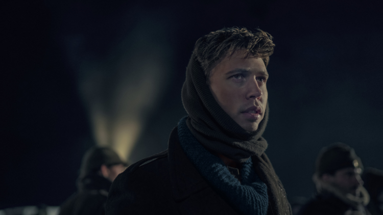 Austin Butler, a white man in his early thirties, looks into the middle distance, stood outdoors at night with a scarf wrapped around his head