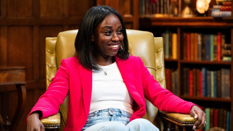 Minah, a black woman wearing a bright pink jacket, sits on a leather armchair smiling