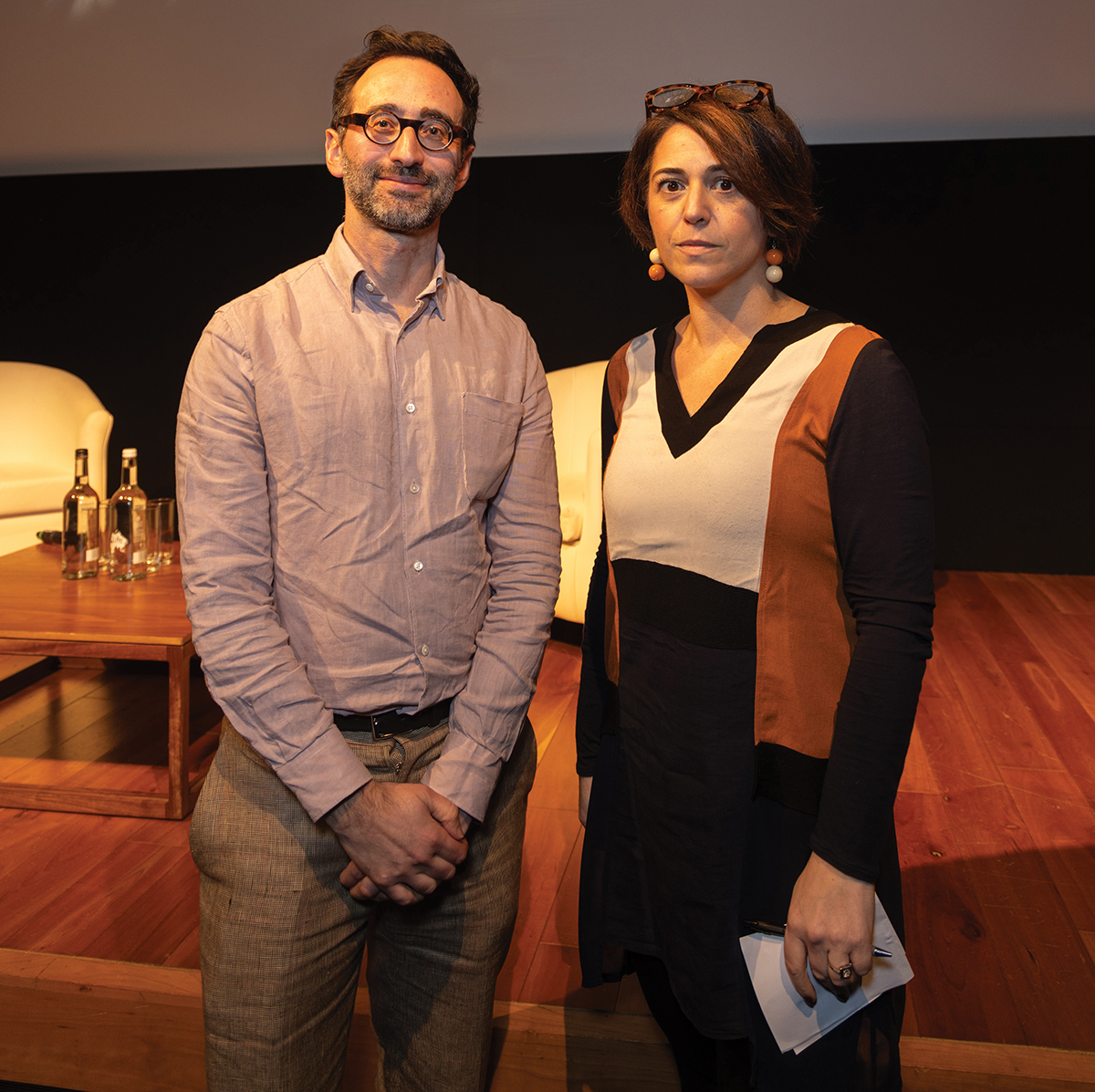 A white man in his 30s with dark hair and glasses, wearing a light pink shirt, stands next to a white woman in her 40s wearing a black skirt and white, black and red top, in front of a stage