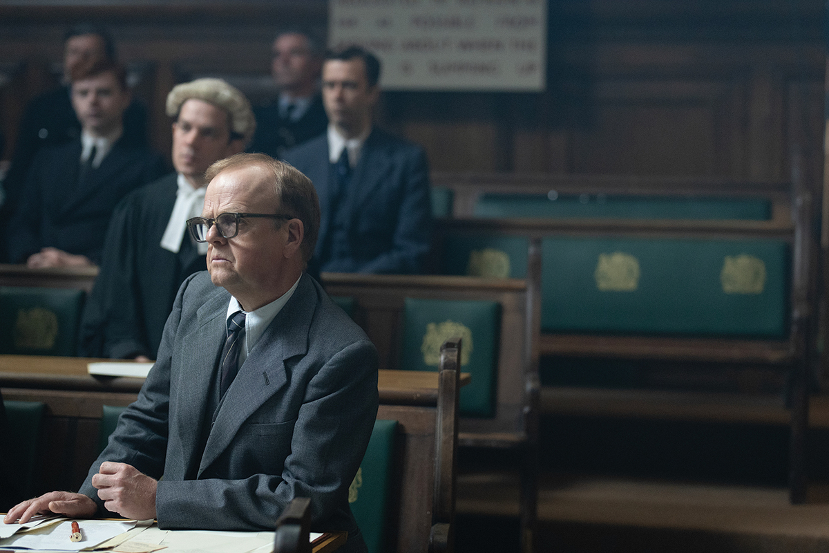 A white lawyer with blond/brown hair stands in a courtroom leaning on a bench