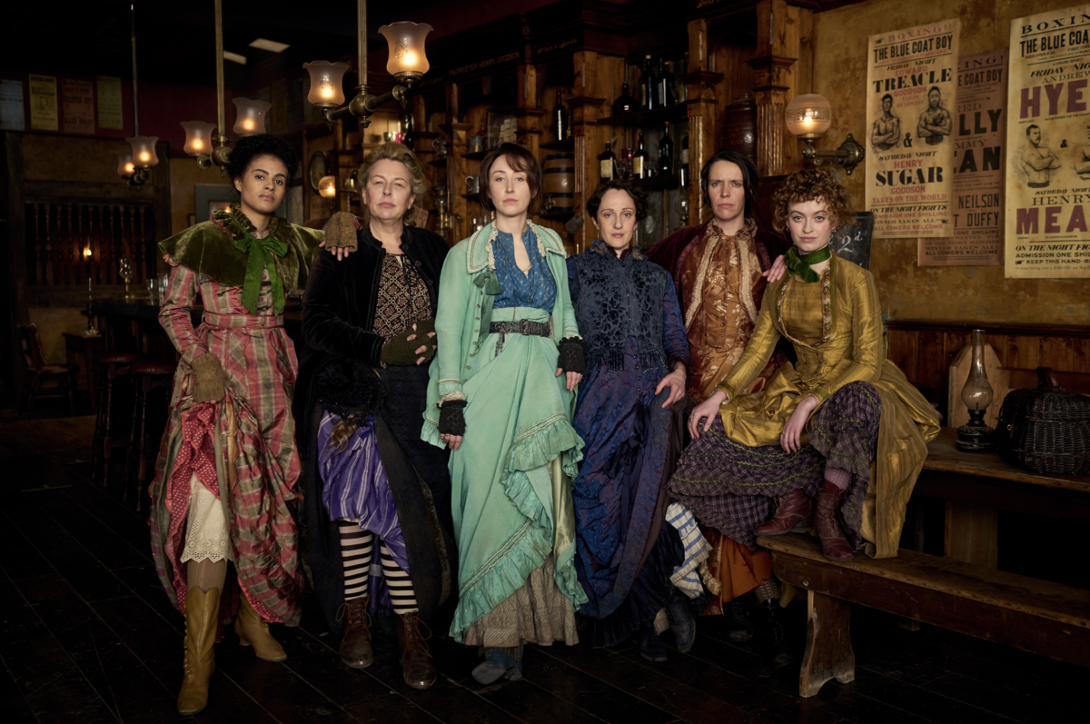 A row of six women of varying ages, skin tones and outfits stand in front of a bar. They are wearing victorian clothing, with some pulling up their skirts to show the boots underneath. There are boxing posters on the walls behind them, 