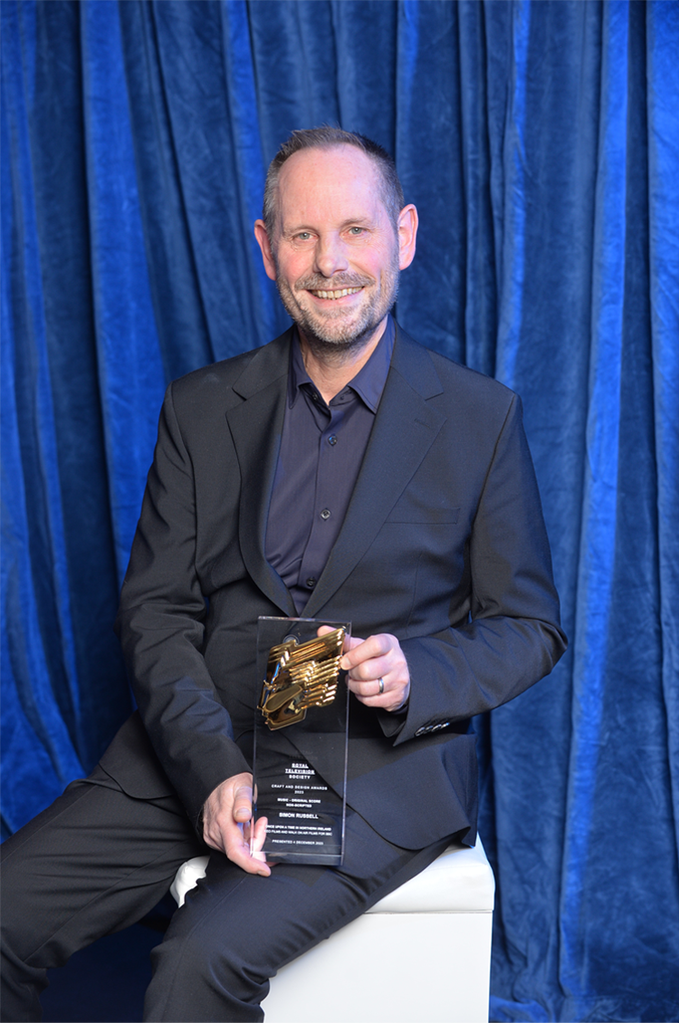 Simon Russell sits in front of a blue backdrop holding his Craft & Design award