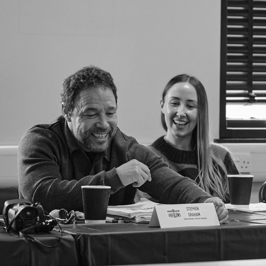 A black and white photo shows Stephen Graham, a man in his early 50s, sat laughing with Erin Doherty, a woman in her early 30s. Both have light skin tones. 