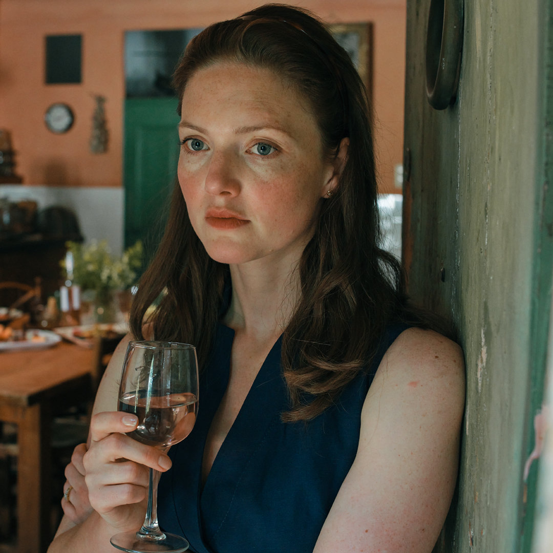 Holliday Grainger, a white woman in her thirties, holds a glass of white wine indoors, wearing a deep blue top 