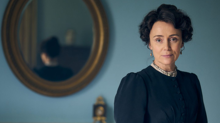 Keeley Hawes, a white woman in her forties, looks into the camera, wearing dark blue 19th century period dress, in front of a wall with a lighter blue hue. She stands to the right, with a mirror hung on the wall to the left