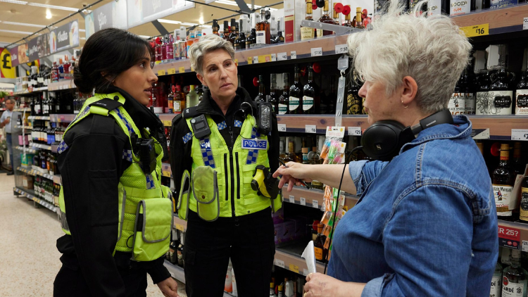 Sally Wainwright gestures at Tamsin Greig and Taj Atwal, who listen attentively, in a supermarket aisle. Greig and Atwal are dressed as police officers, including hi-vis tabards, while Wainwright wears a blue denim shirt and black over-ear headphones