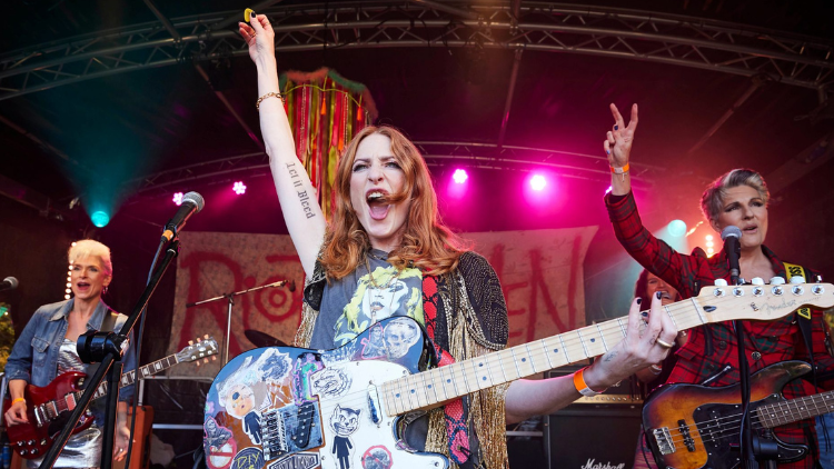 Amelia Bullmore, Rosalie Craig, Macy-Jacob Seelochan and Tamsin Greig stand onstage at an outdoor festival, Craig’s arm raised – holding a guitar pick – mid-shouting, and Greig’s arm raised, giving a peace symbol