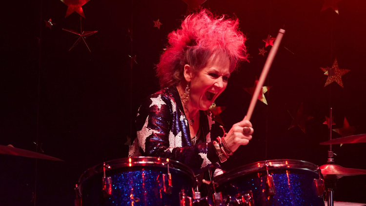 Lorraine Ashbourne, a white woman in her 60s, shouts as she plays the drums, wearing a sequined top with white stars and basked in pinkish red light