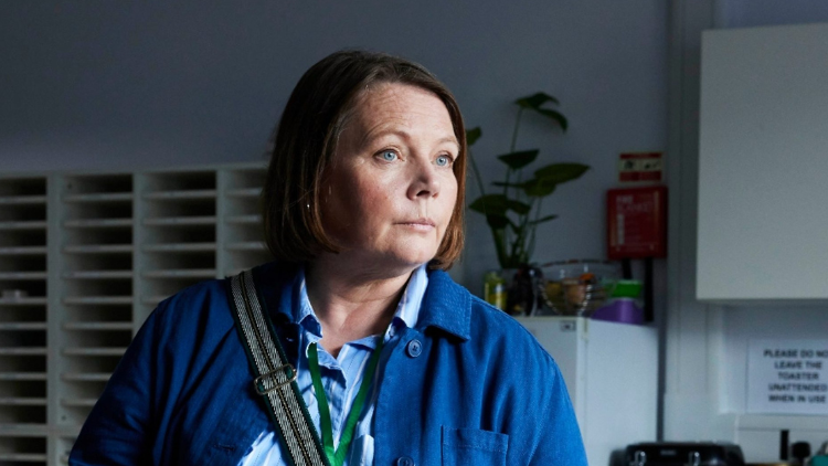 Joanna Scanlan, a white woman in her sixties, looks at something off-camera, wearing a blue jacket and light blue shirt, stood indoors