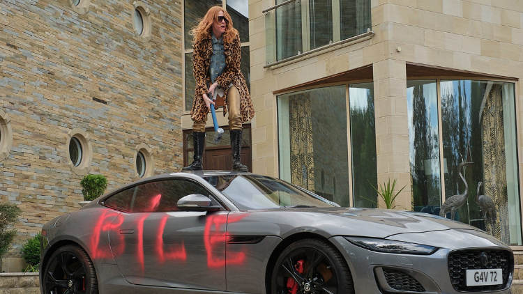 Rosalie Craig, a white woman in her forties, stands on top of a grey sports car with the word ‘bile’ spraypainted in pink on the side. She is holding a large, metal mallet, and wears a leopard print jacket